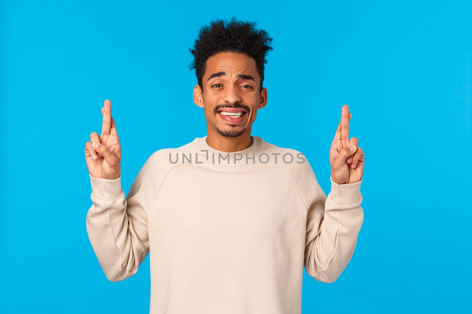 Nervous timid and cute african-american guy with afro haircut scared telling parents something huge, pointing fingers up, cringe and smiling with anxious expression, feeling alarmed, blue background by Benzoix