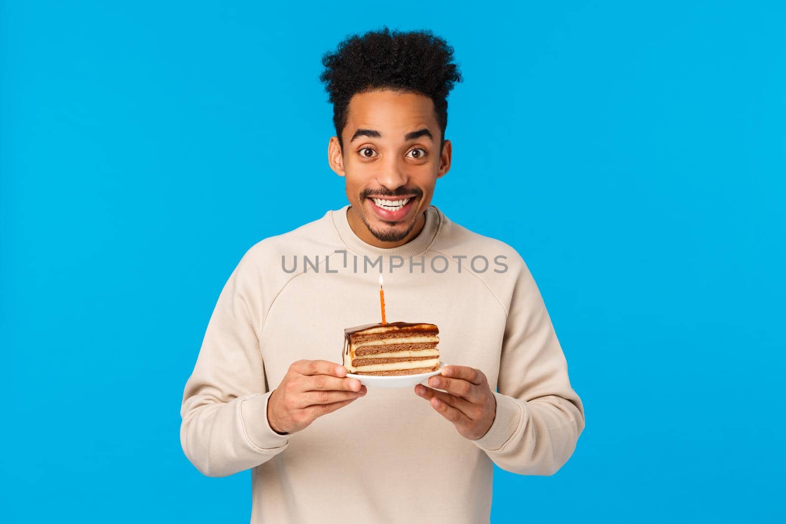 Cheerful and excited happy smiling african american b-day guy celebrating birthday, holding piece cake with candle, making wish ancitipating good year, standing blue background.