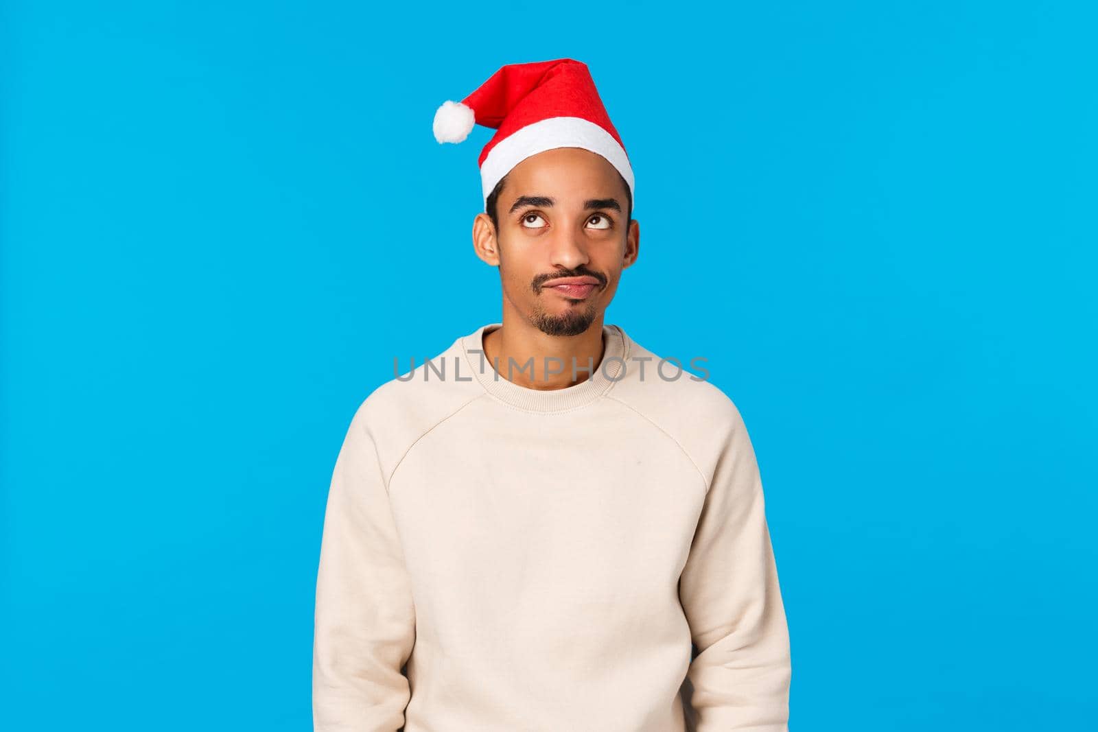 Worst new year ever. Upset and distressed young skeptical african-american guy dont feel christmas mood, standing sad and disappointed, wearing santa hat and winter sweater, blue background.