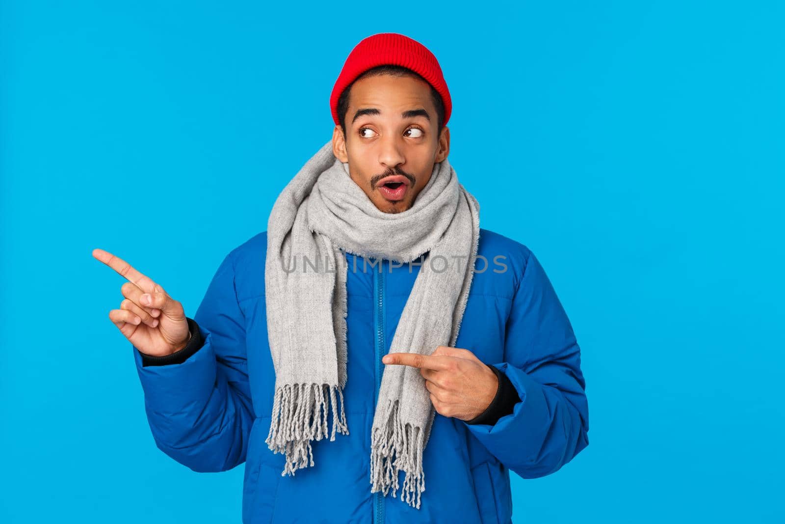 Intrigued and curious good-looking african american young hipster guy snowboarder looking at awesome huge snowy cliff to slide from, pointing upper left corner say wow astonished, blue background.