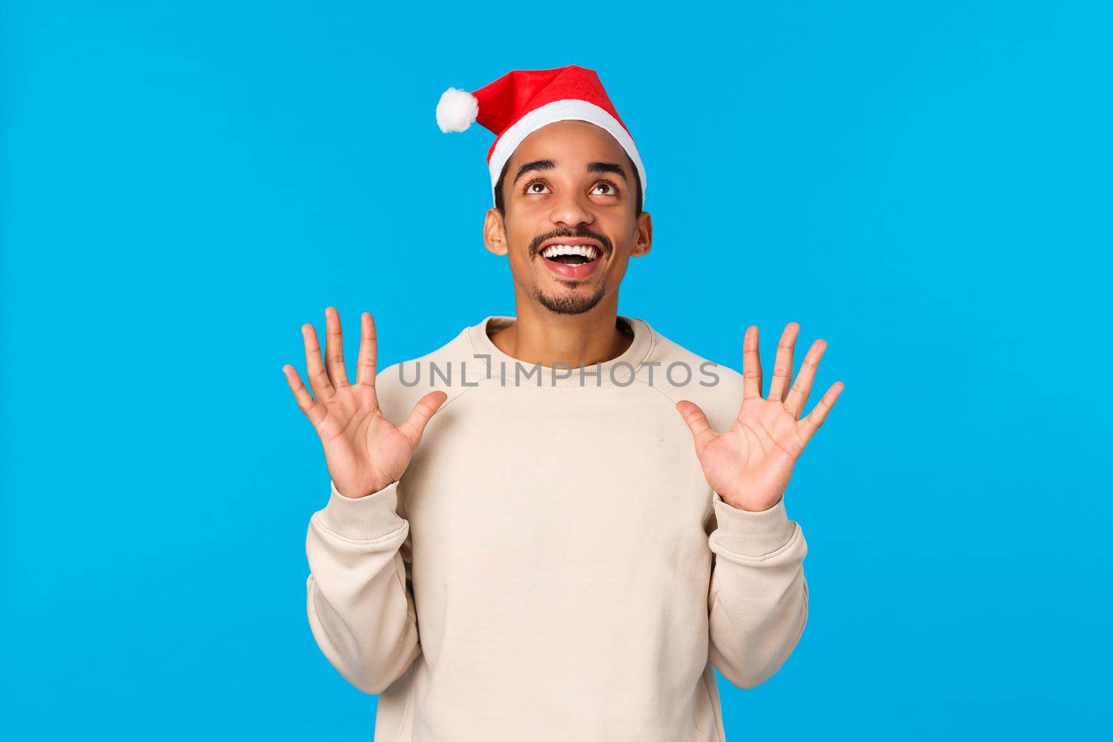 Celebration, christmas mood and happiness concept. Cheerful and happy african-american young man having fun, wearing santa hat and winter sweater, catching gifts from upwards, smiling look up.