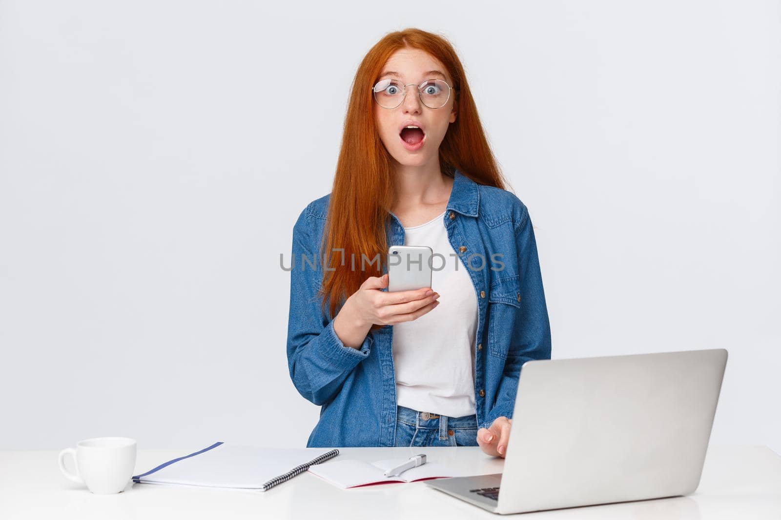 Amazed, surprised and shook teenage redhead girl staring speechless camera, drop jaw gasping wondered look, read fascinating news, got message on smartphone, stand near laptop and desk.