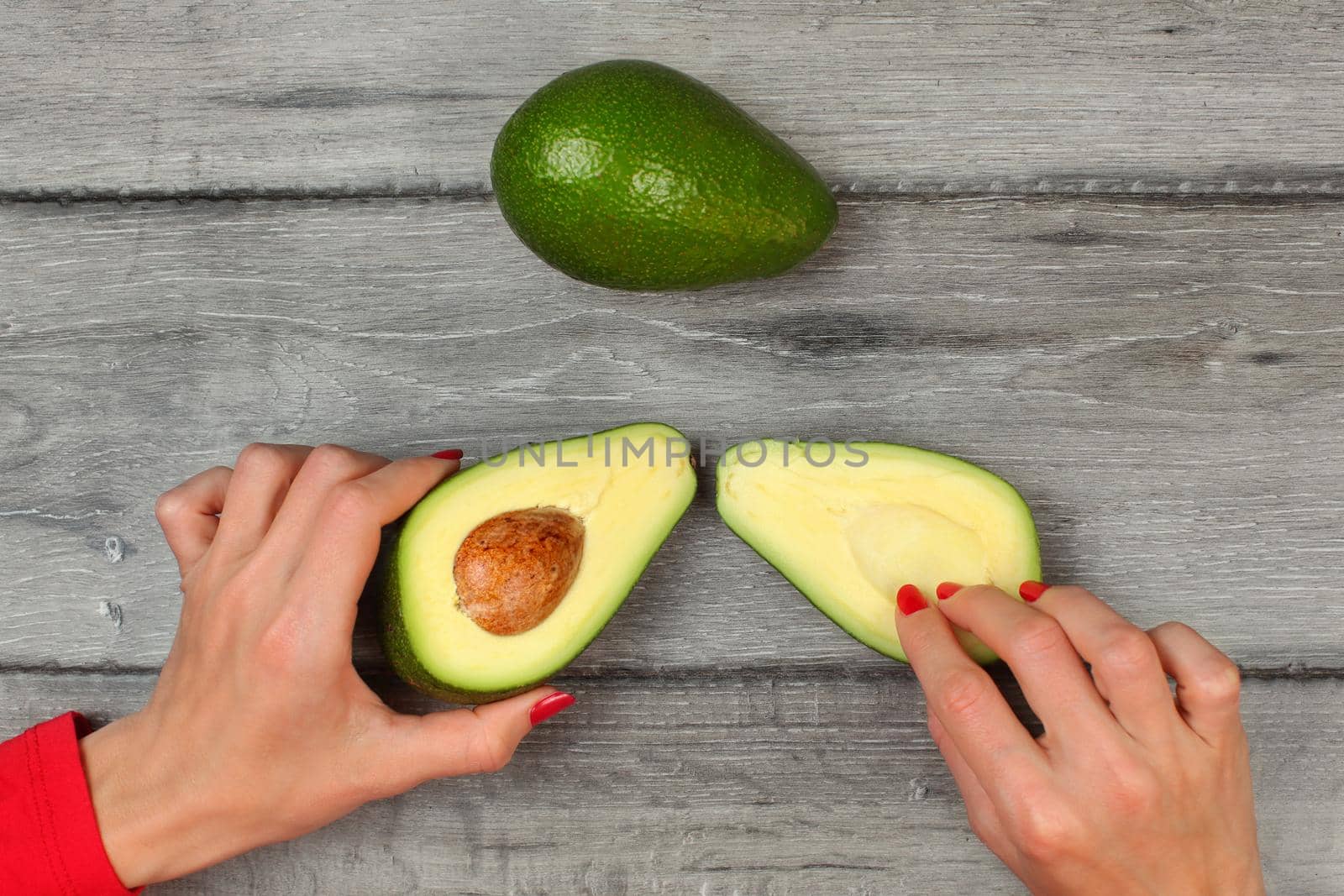 Table top view - woman hands holding avocado cut in half, another one above, placed on gray wood desk