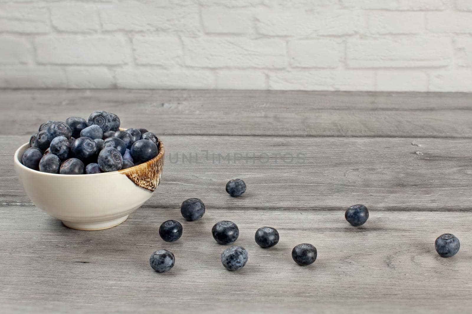 Small bowl full of blueberries, some of them spilled on gray wood desk.