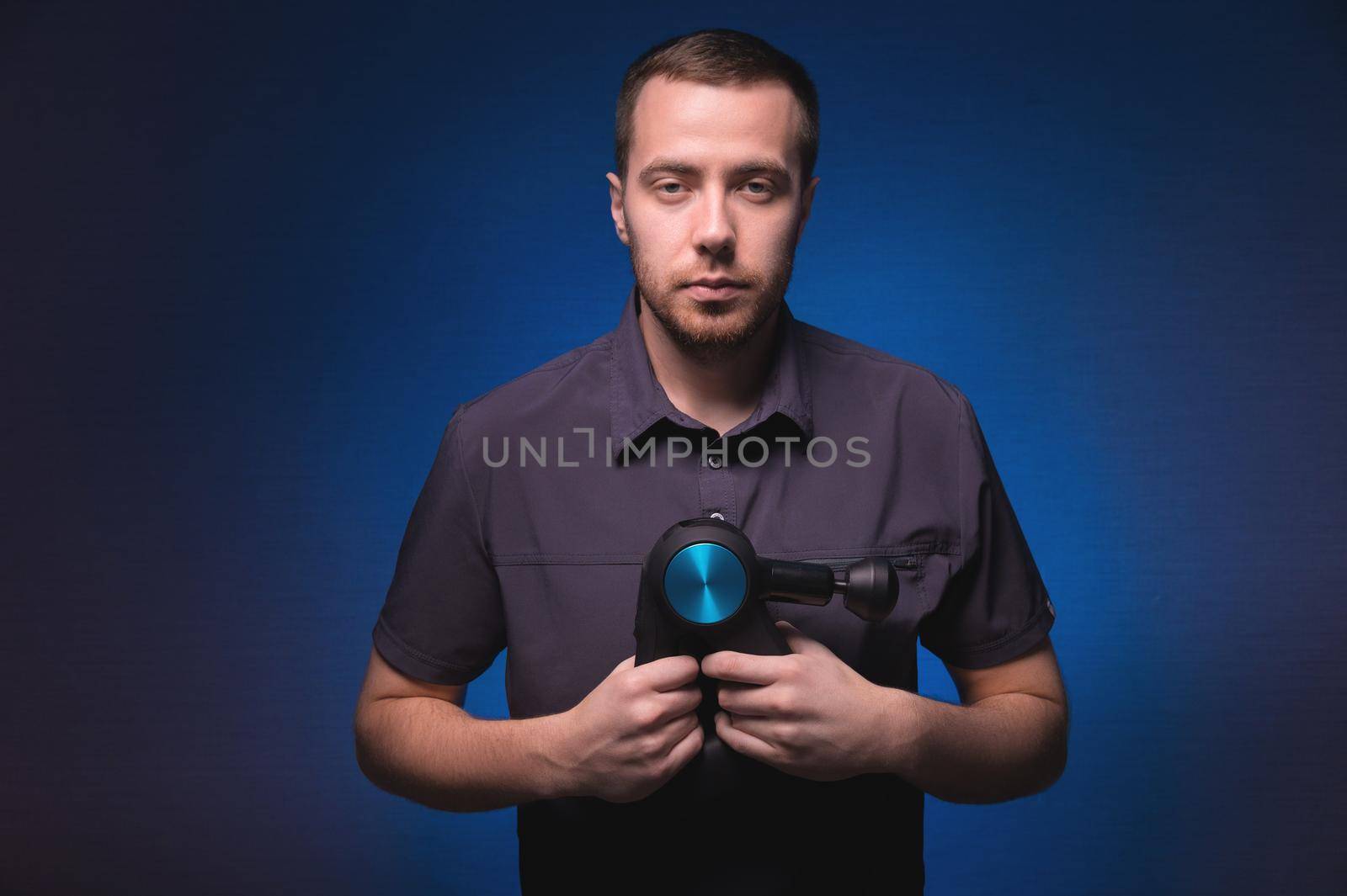Portrait of a confident professional masseur with a percussion massager in his hands. Low key, blue background. Shock wave massage by yanik88