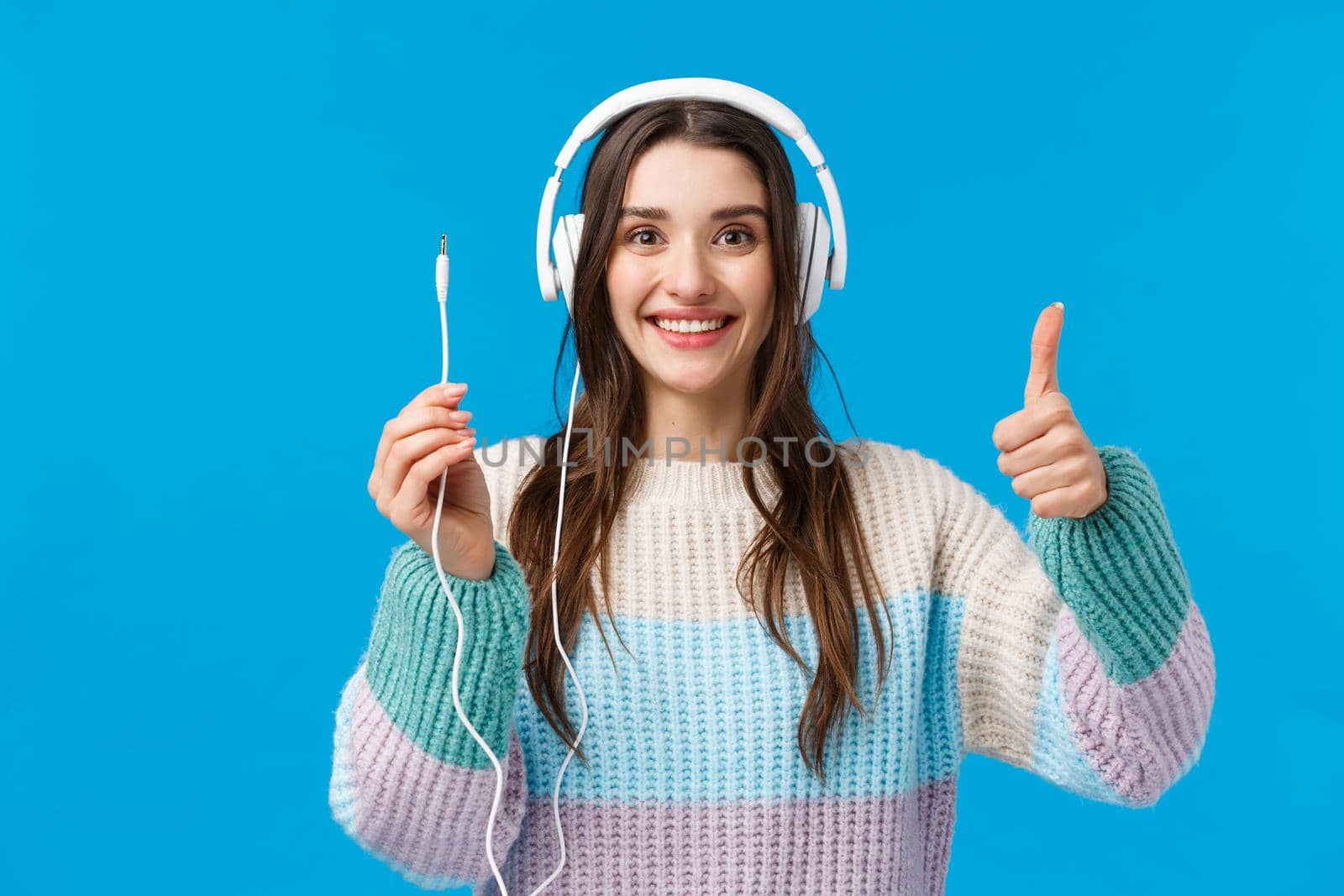 Waist-up portrait cheerful, excited young woman smiling, showing thumb-up in approval, like as received awesome present, wearing new headphones, holding wire and grinning.