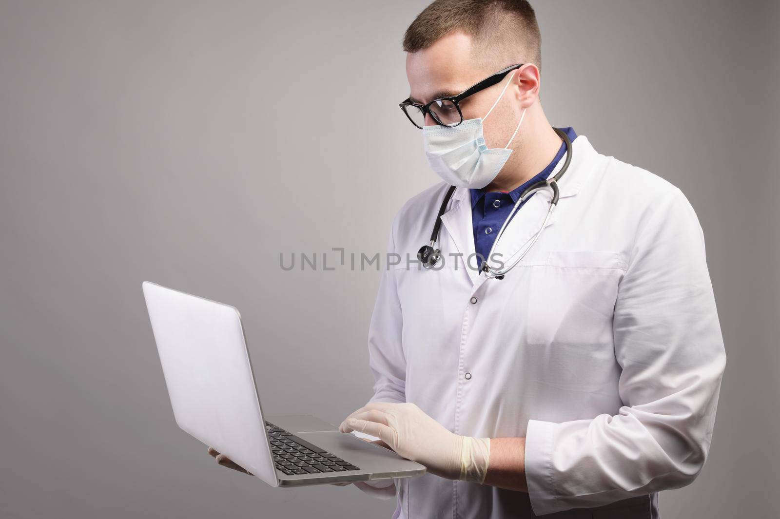 Successful young caucasian male doctor in a white medical uniform and with a stethoscope, busy working on a laptop, typing on a computer, consulting a patient online. Studio portrait.