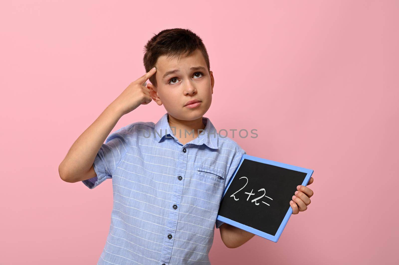 A schoolboy with a blackboard in his hand holds a finger to his temple and with a thoughtful look solves an arithmetic task, standing on a pink background with a place for text. Back to school. Concepts