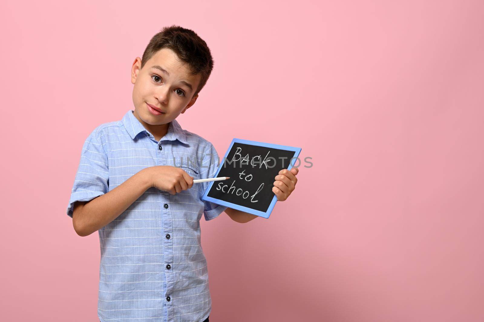 Smiling school boy holding a chalkboard with lettering Back To School and pointing with a pencil on it, posing over pink background with space for text. Concepts