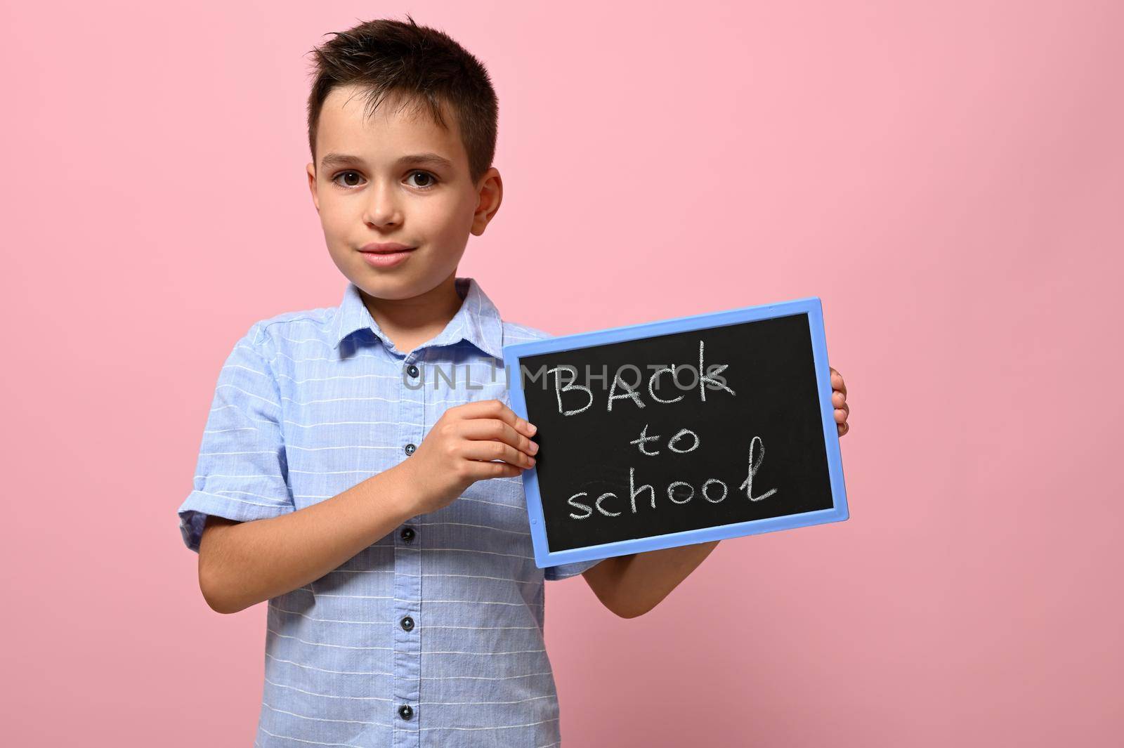 An adorable schoolboy holding a chalkboard with chalk lettering ,Back to school, isolated over pink background with space for text