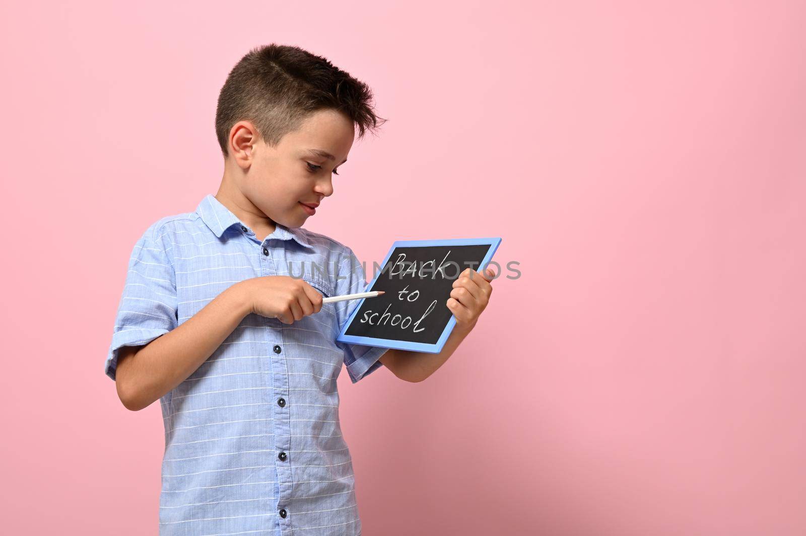 Lettering Back To School on a chalk board in the hands of a happy pupil. Isolated over pink background with copy space by artgf