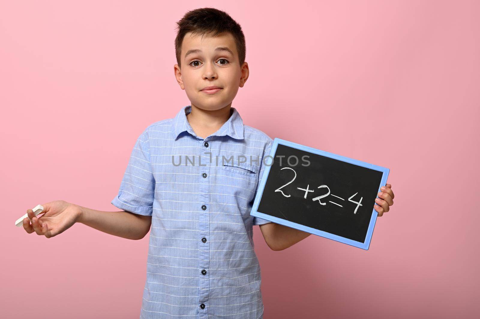 Adorable boy, elementary school student holds chalk and blackboard and solves math problem. Pink background space for text. Back to school. Concepts by artgf