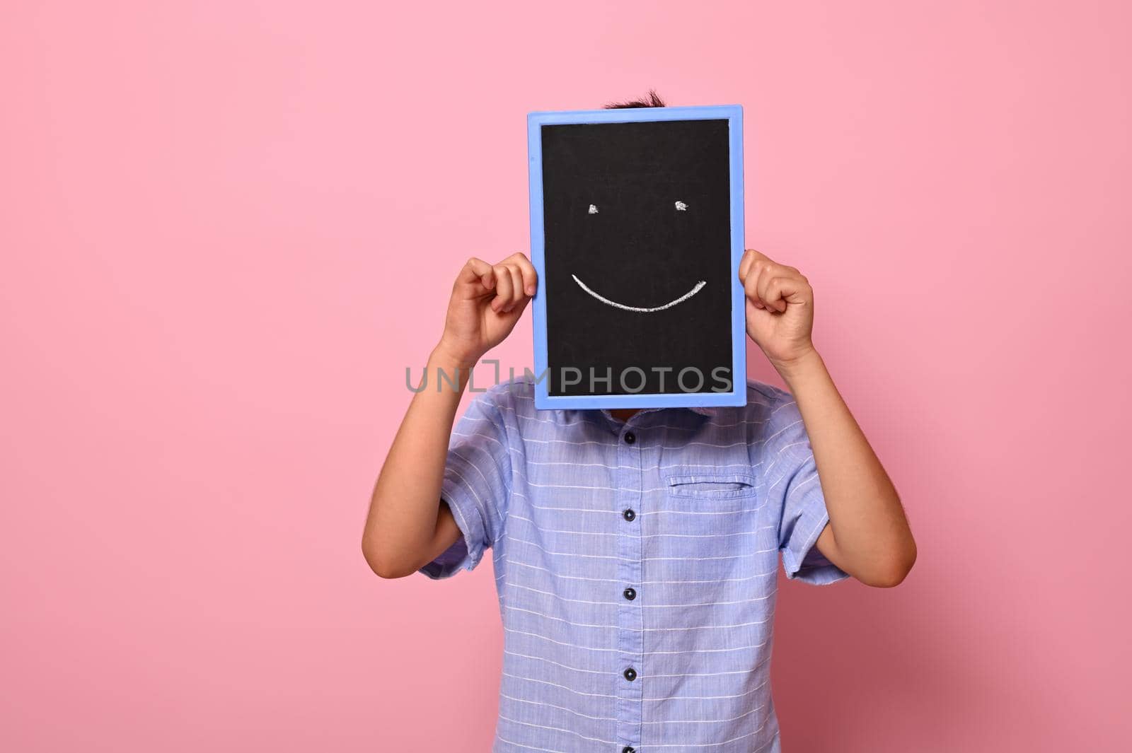 A school boy covers his face with a chalkboard with drawn smiling emoticons, expressing happiness. Isolated over pink background with copy space by artgf