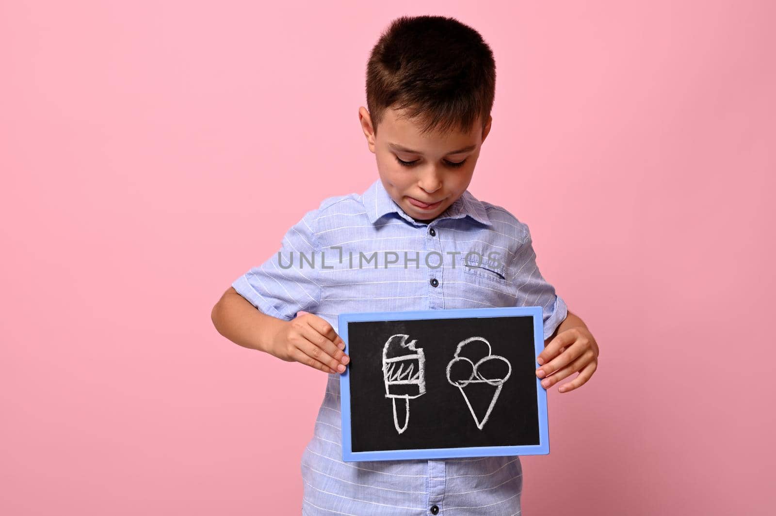 A boy looking at a chalkboard in his hand with drawn ice cream against pink background with copy space. concepts by artgf