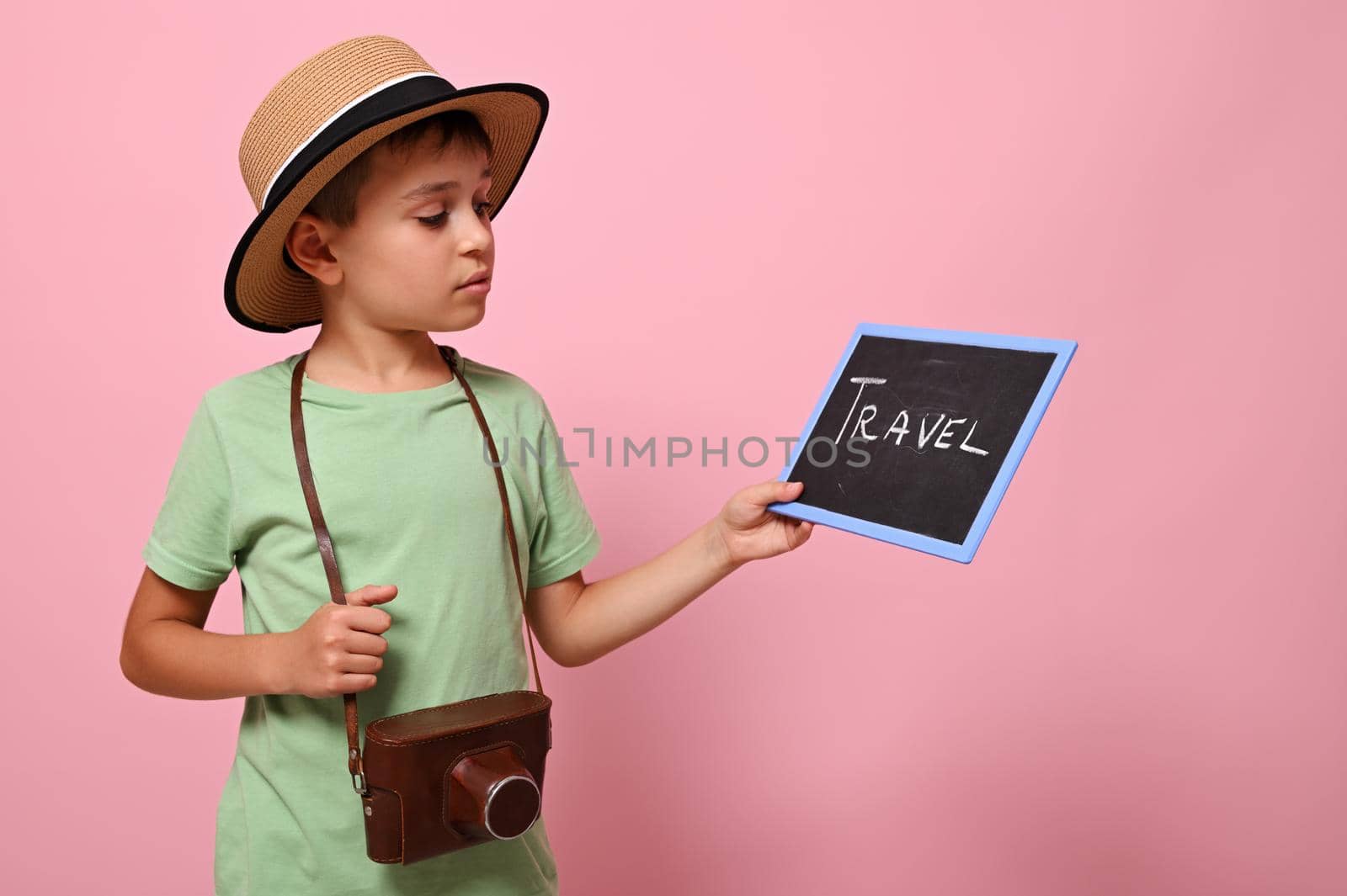 Handsome boy in summer hat with a vintage camera around the neck looking at blackboard with lettering TRAVEL . Tourism and summer concepts on a pink background with copy space by artgf