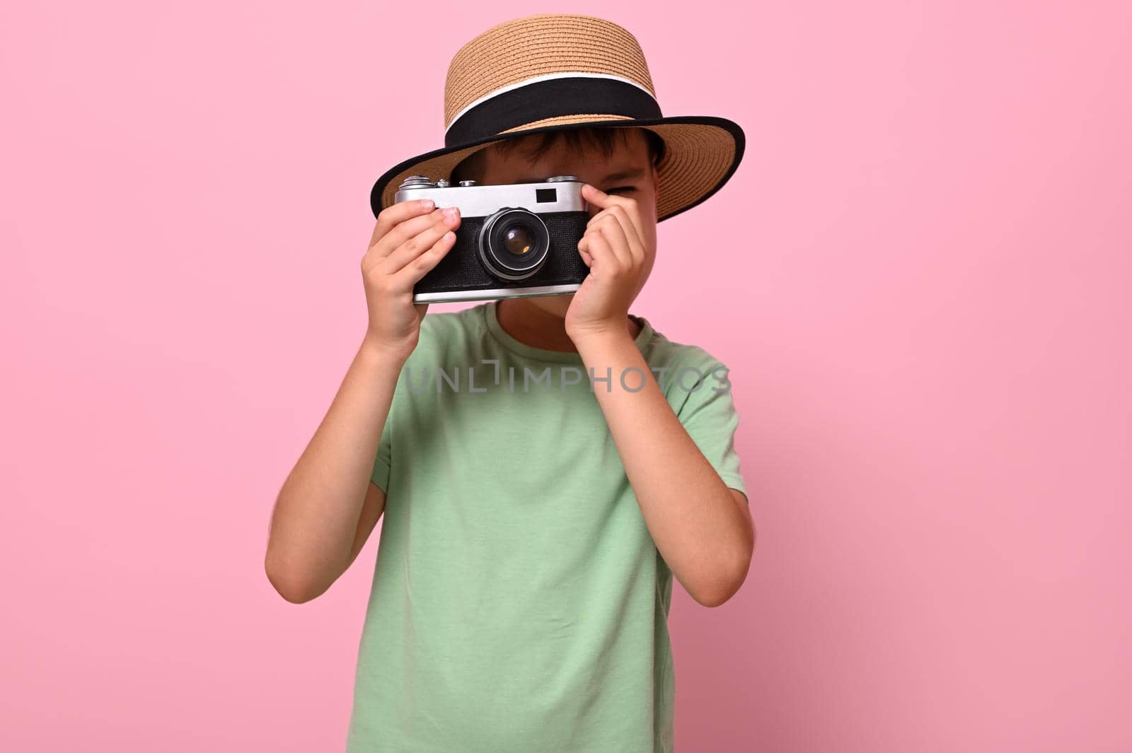 Cute schoolboy in green t-shirt and summer hat taking photo using an old vintage retro camera, isolated over pink background with copy space