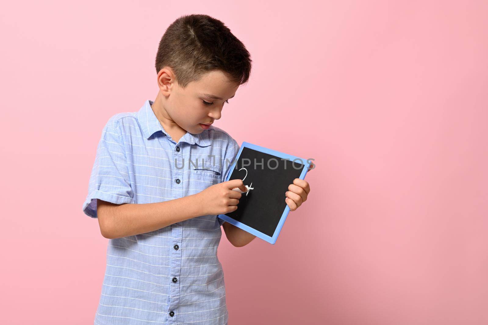 A school boy in blue shirt writes with chalk on a blackboard, isolated over pink background with copy space. Back to school, concepts by artgf