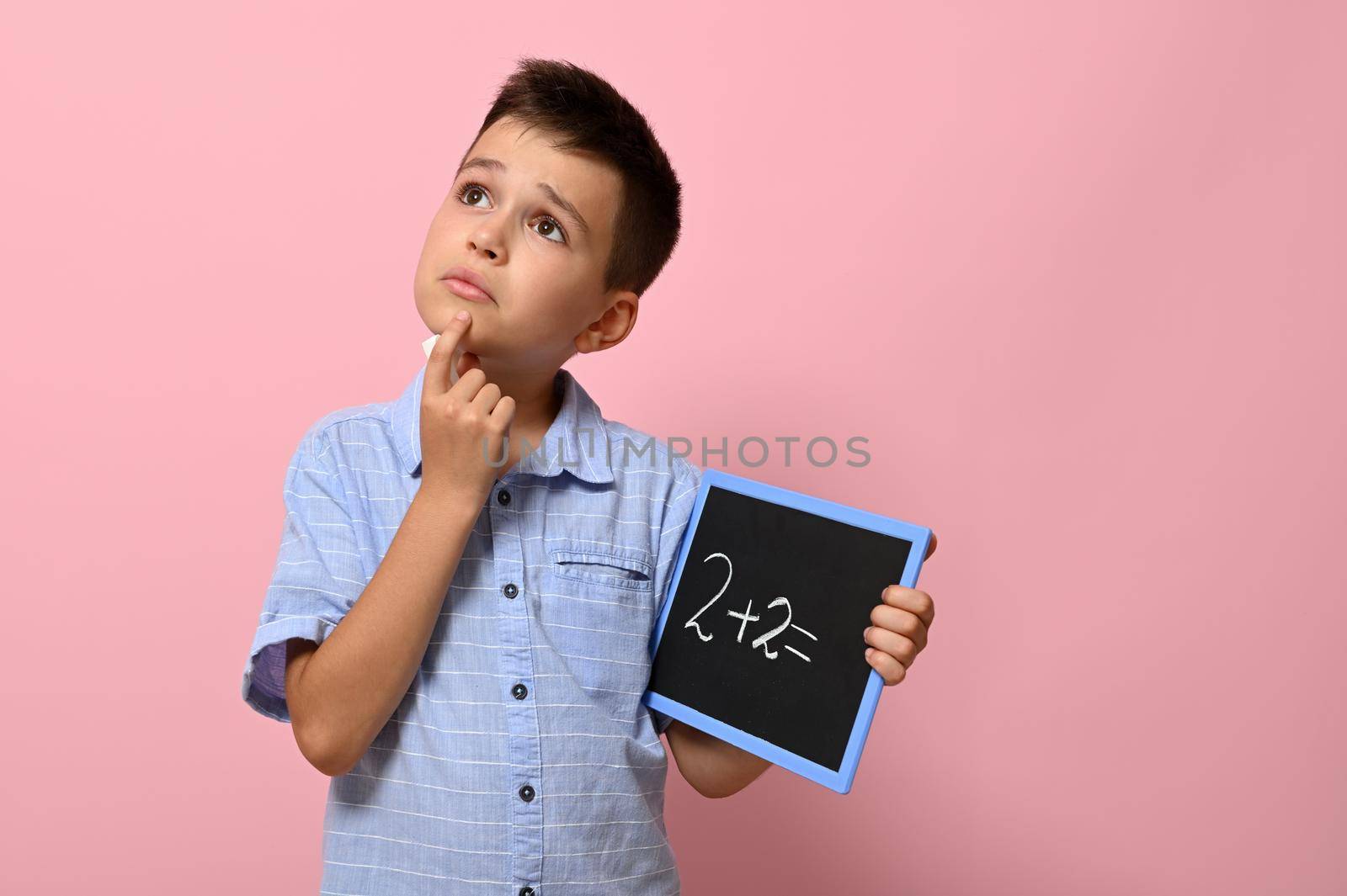 Cute schoolboy holding a chalkboard with chalk writing on it, thoughtfully solves a math problem. Isolated on pink background with copy space by artgf