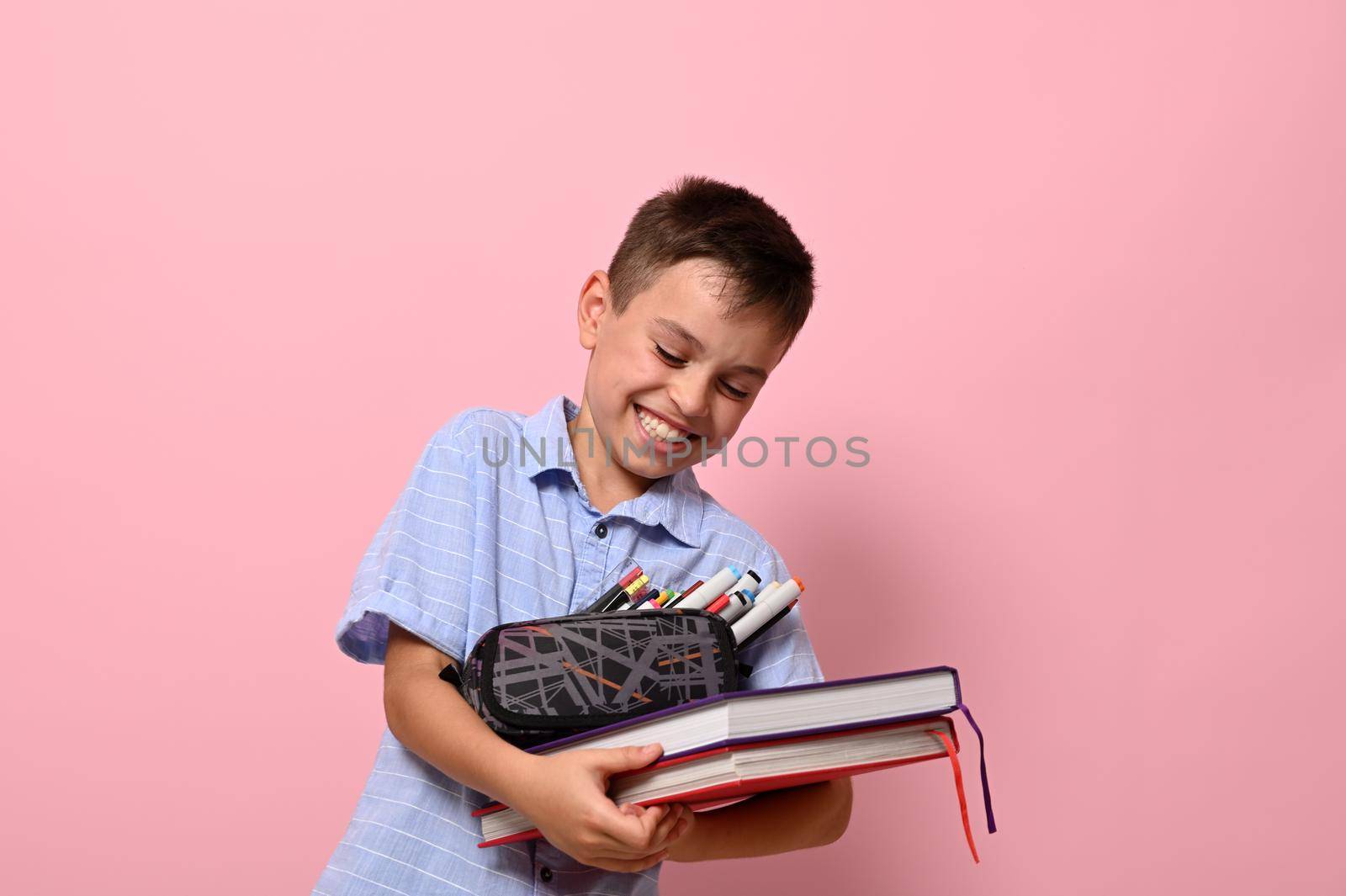 Adorable school boy with pencil case and books laughing , posing over pink background with copy space. Concepts of back to school with facial expressions and emotions