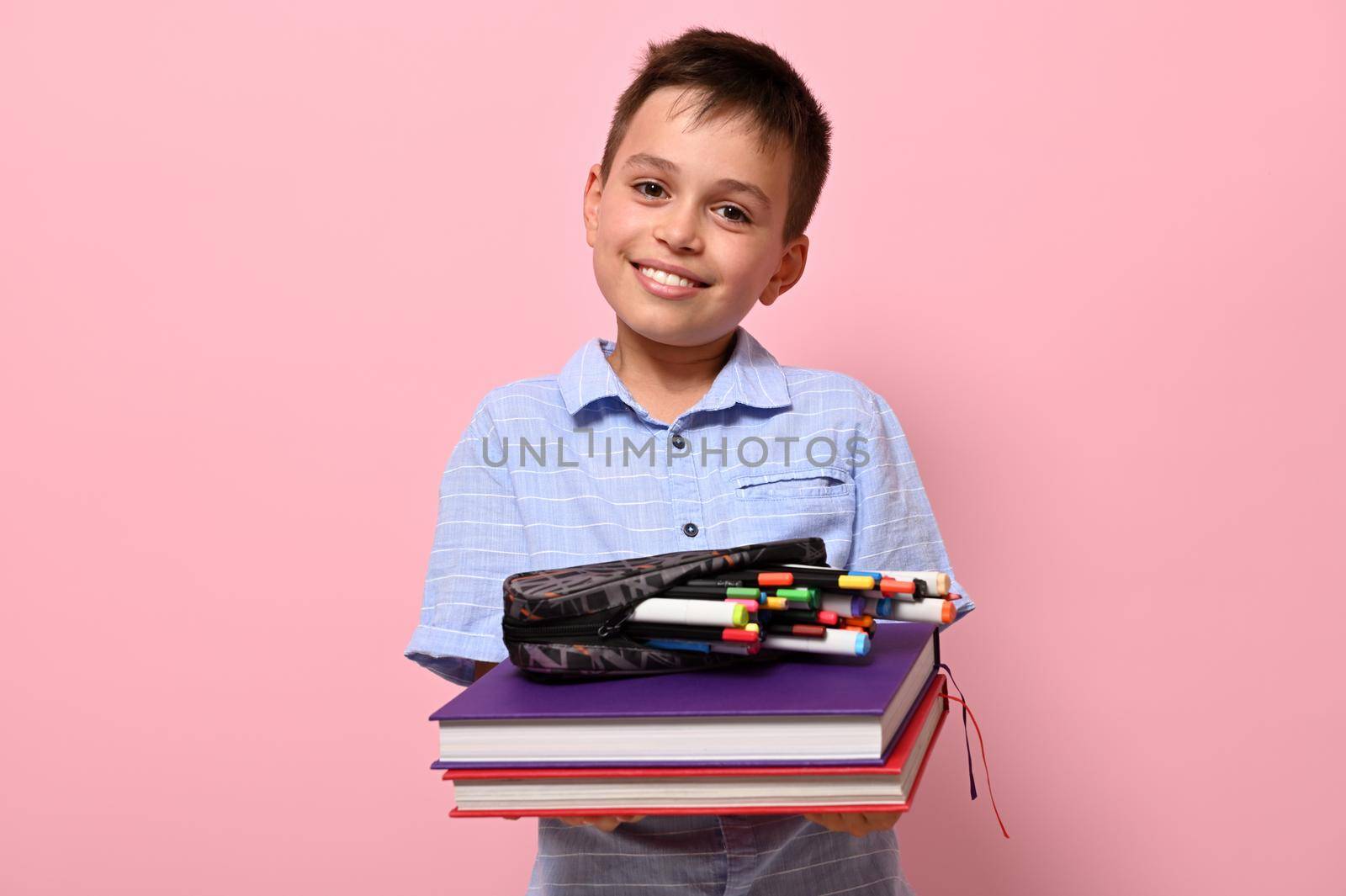 A smiling student at school, holds books in front of him and pencil case with pens, felt-tip pens and markers falling out of him. Back to school concepts on pink background with copy space by artgf