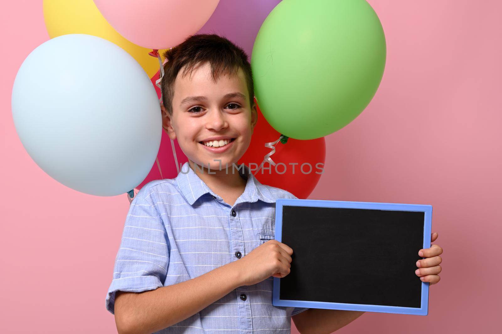 Adorable smiling schoolboy with empty blank chalkboard in his hand standing against multicolored balloons on pink background with copy space by artgf