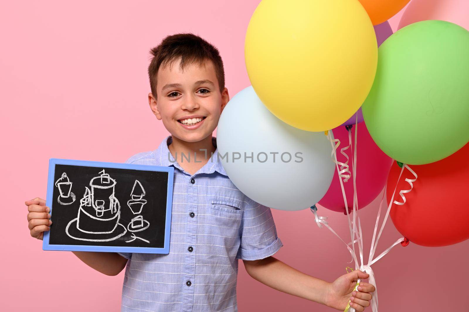 Smiling teenage boy holds multicolored colorful balloons in one hand and a chalkboard with drawn birthday cake in the other. Isolated over pink background with copy space