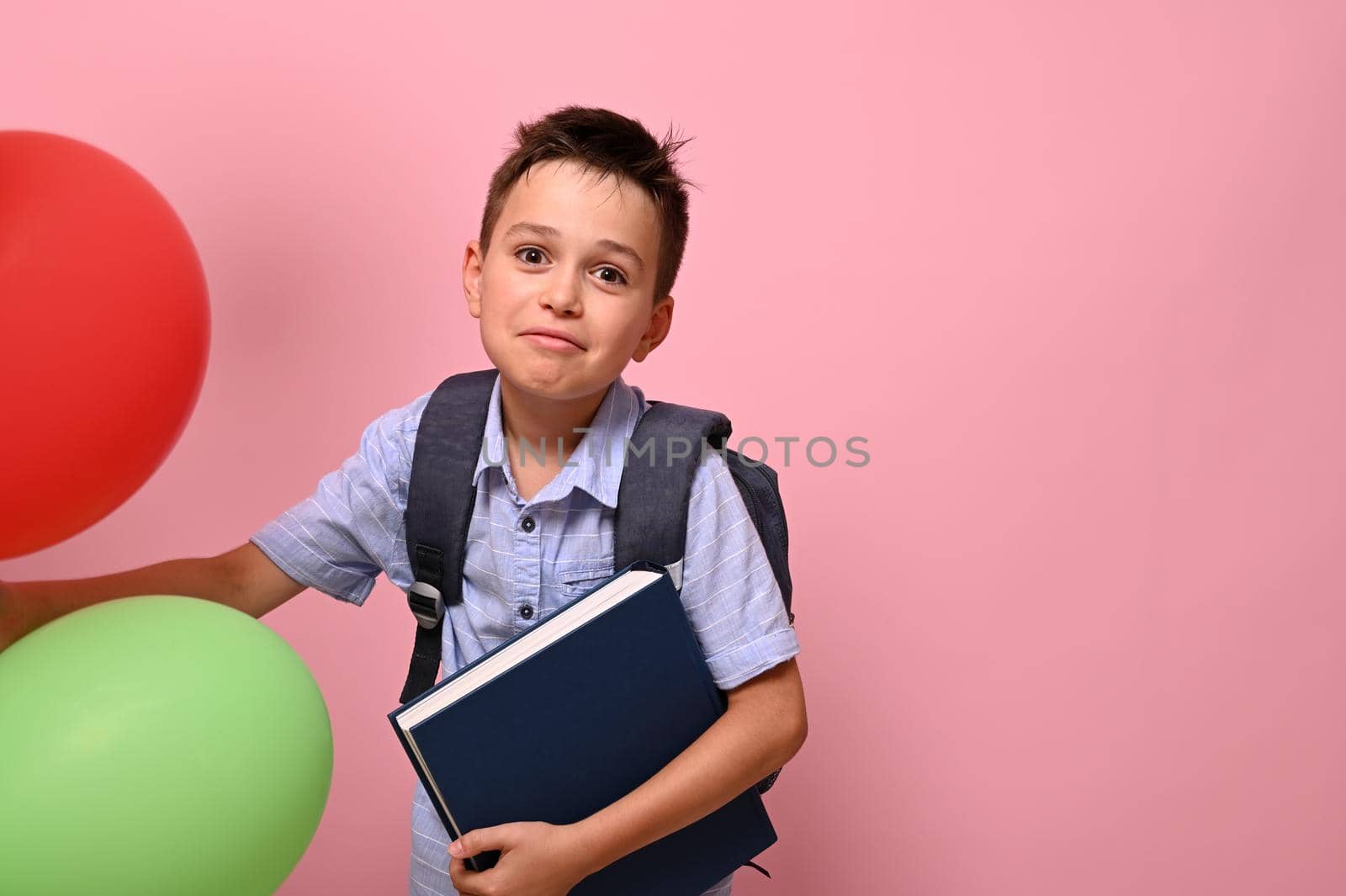 Concepts of happy back to school. Schoolboy with backpack holding book and multicolored balloons, cute smiling posing to camera over pink background with copy space by artgf