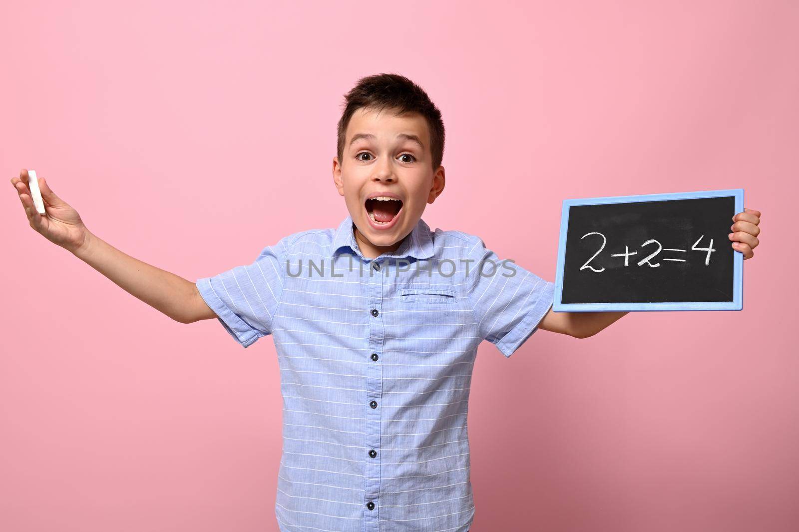 An adorable boy, an elementary school student, holds a chalk and a chalkboard and happily solves math problems. Pink background for text by artgf