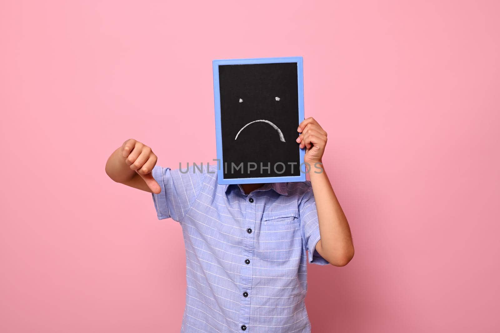 A school boy covers his face with a chalkboard with drawn smiling emoticons, expressing sadness and shows thumb down to the camera. Isolated over pink background with copy space