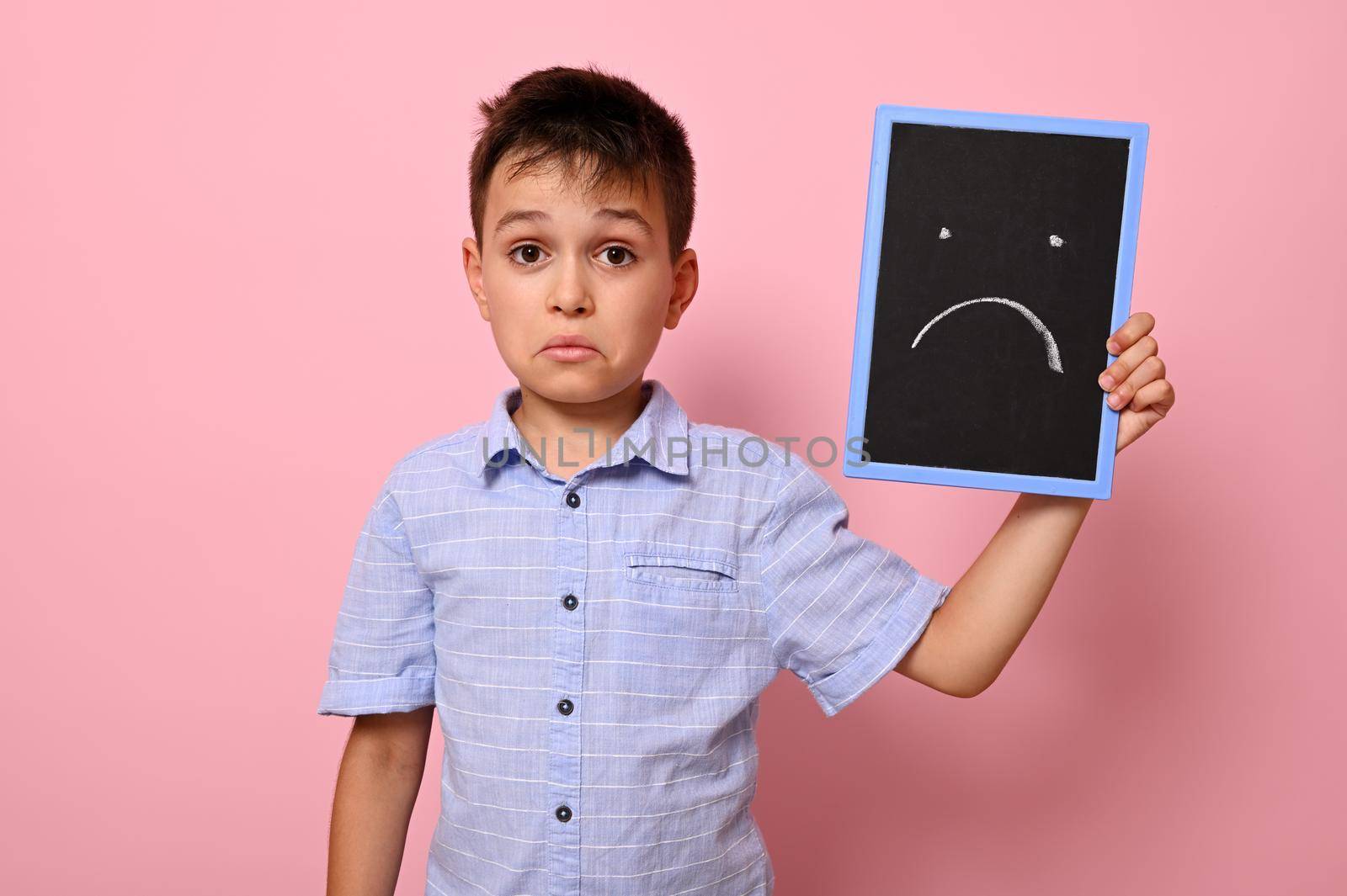 A frustrated boy in a blue shirt holds a blackboard near his face with a painted drawn expressing frustration, sadness and disappointment. Pink background, copy space by artgf