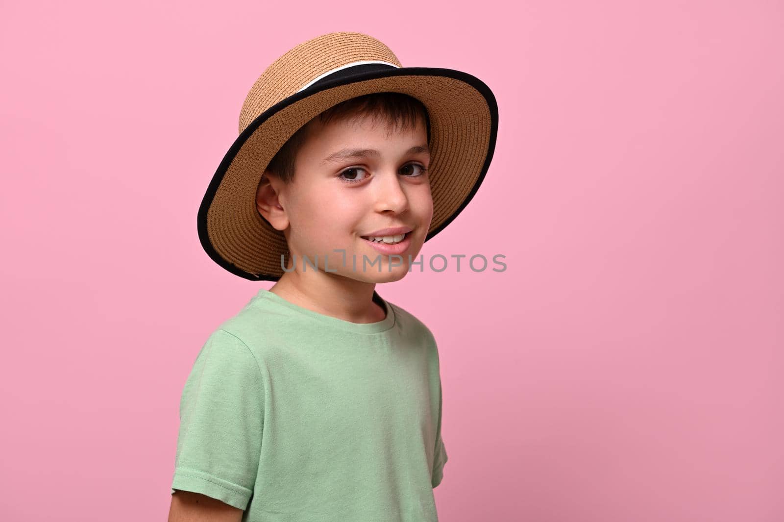 Side portrait of a handsome schoolboy in summer straw hat, looking at camera and standing isolated over pink background with copy space. Close-up, headshot.