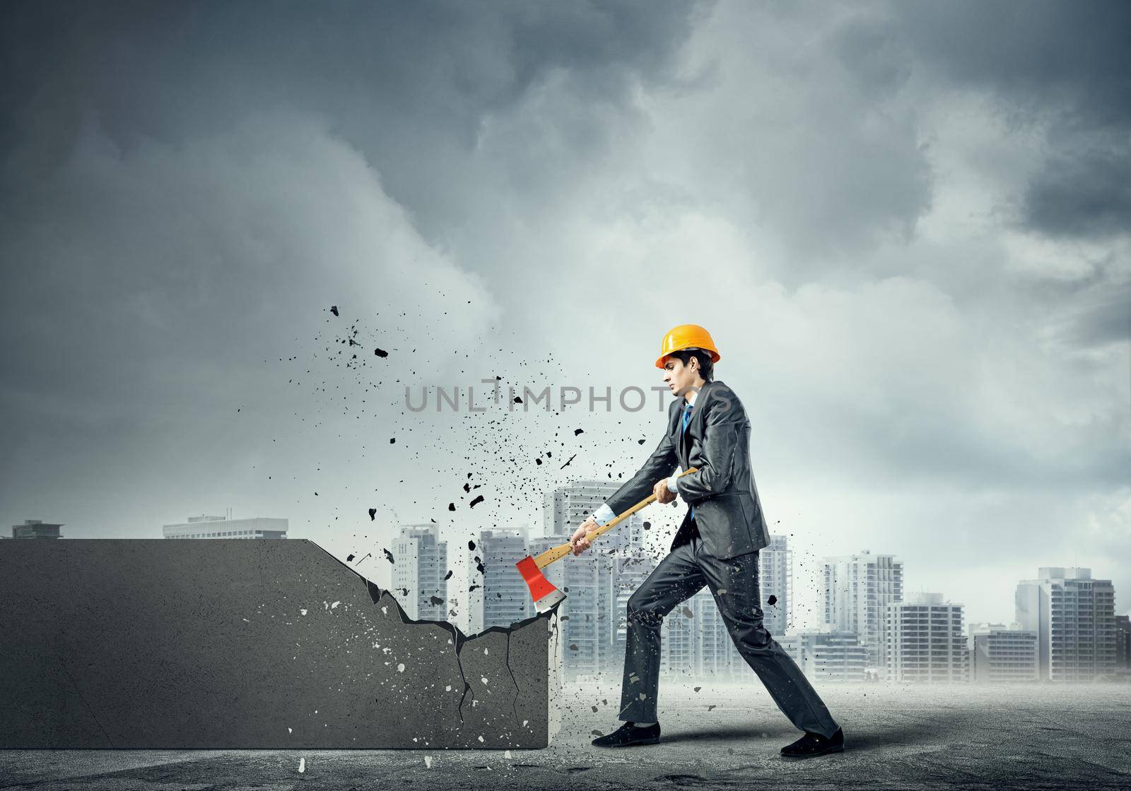 Young businessman in helmet breaking cement wall with axe