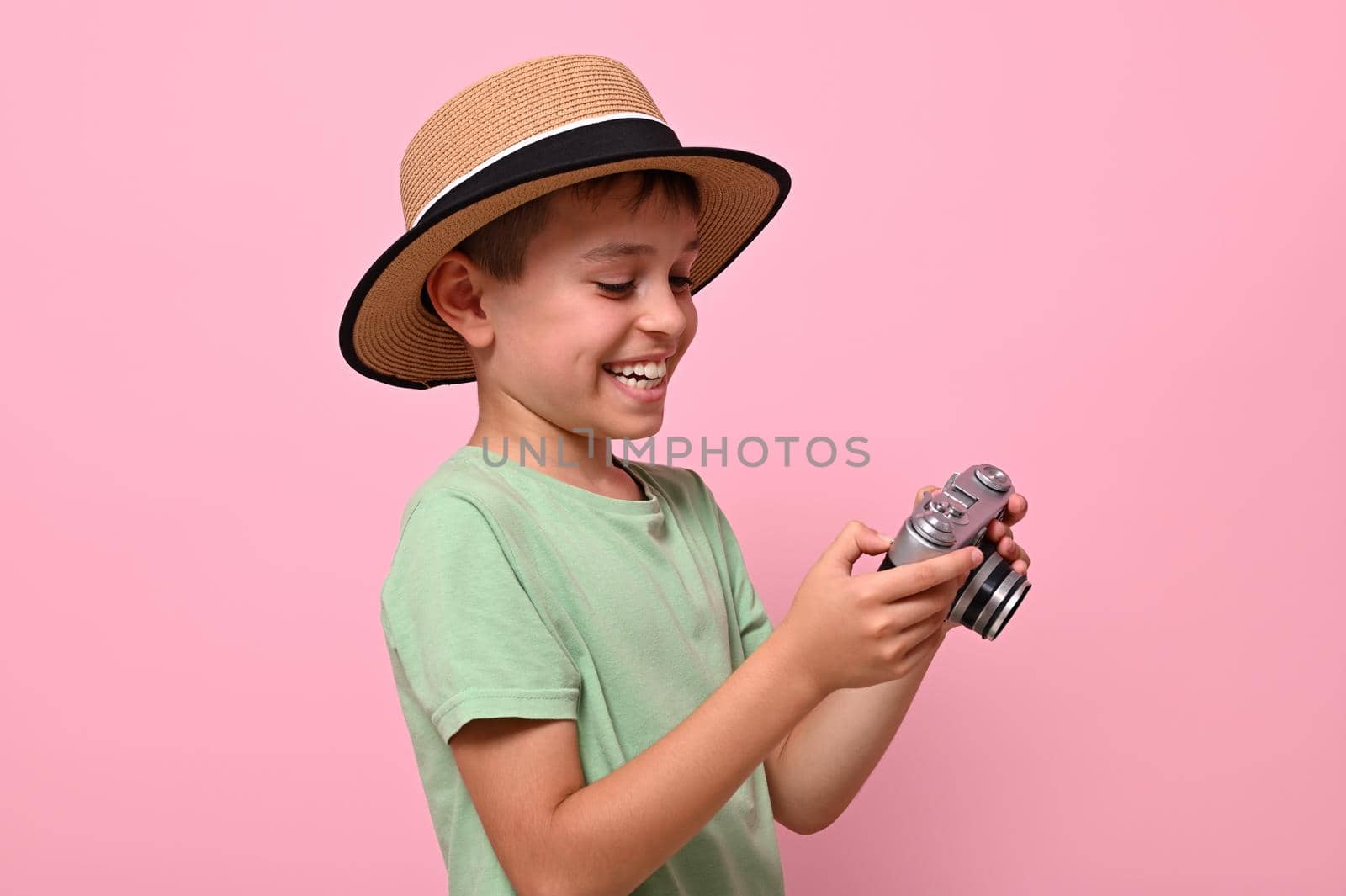Happy child in summer outfit holding a retro vintage camera and cutely smiles with toothy smile posing over pink background with copy space. Tourism concepts by artgf