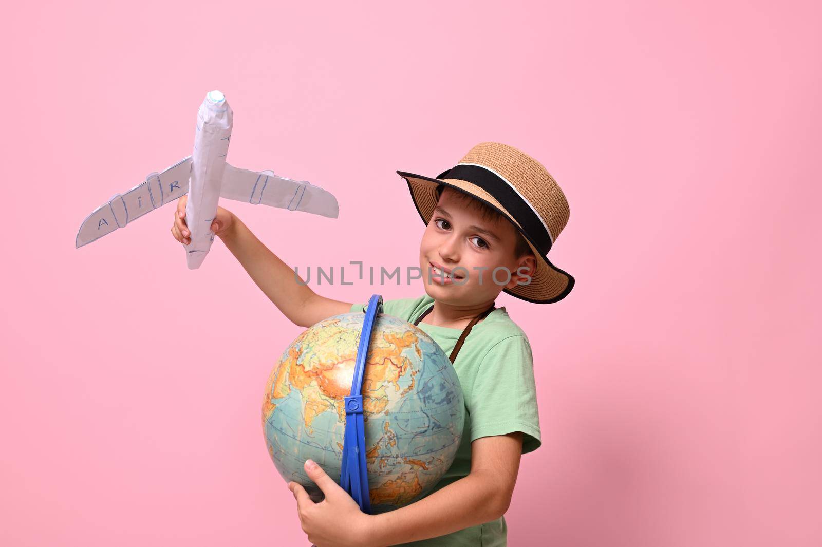Smiling school boy, tourist, looking at camera while playing with paper airplane and a globe, standing isolated over pink background with copy space. Tourism, travel, geography knowledge concepts