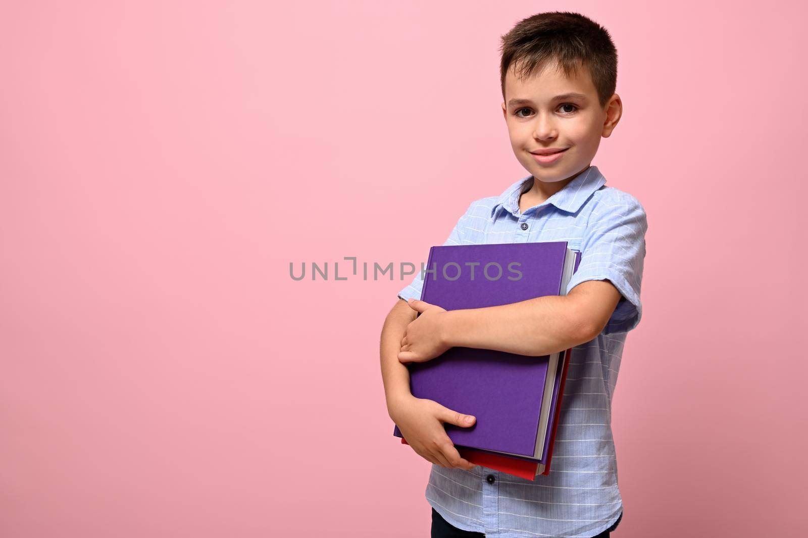 An adorable schoolboy holds books in his hands a cute smiles posing on camera, isolated over pink background with space for text. Back to school concepts