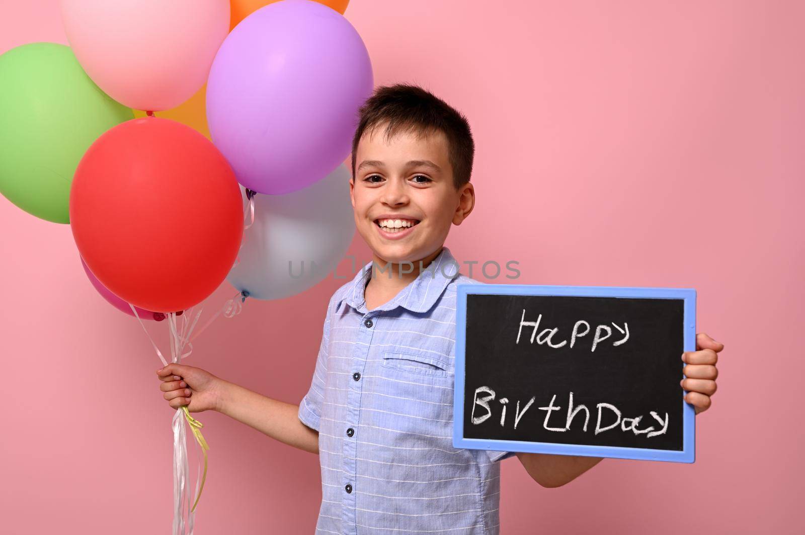 Happy and smiling teenage boy holds multicolored colorful balloons in one hand and a chalkboard with lettering Happy Birthday in the other. Isolated over pink background with copy space