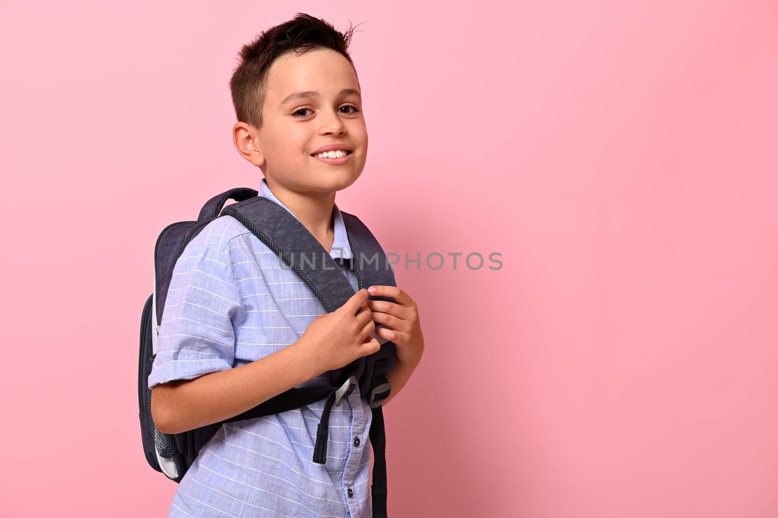 Side portrait of a smiling with toothy smile schoolboy with a backpack on his back on pink background with copy space. Back to school concepts