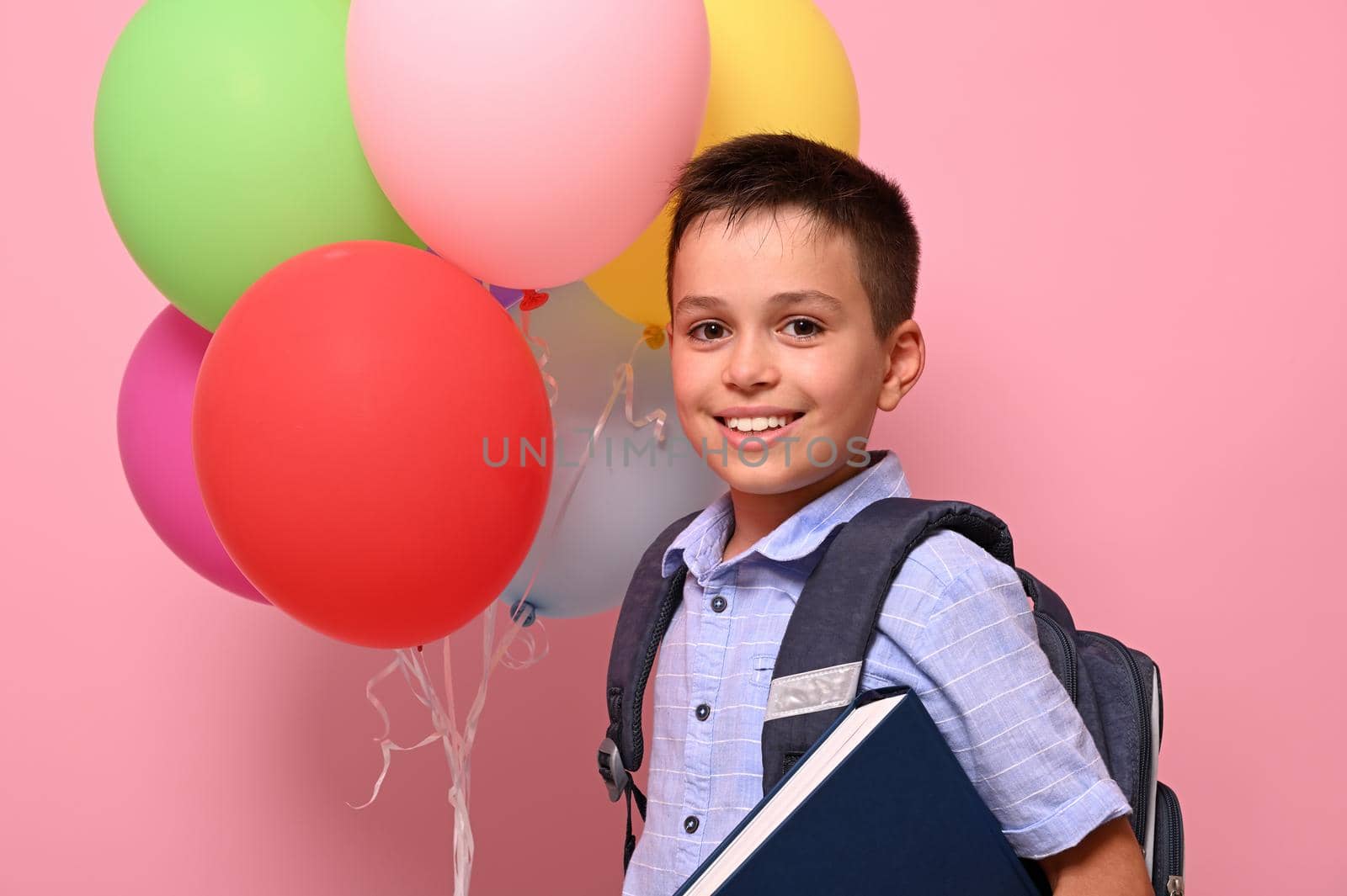 Concepts of happy back to school. Schoolboy with backpack holding book and multicolored balloons, cute smiling posing to camera over pink background with copy space