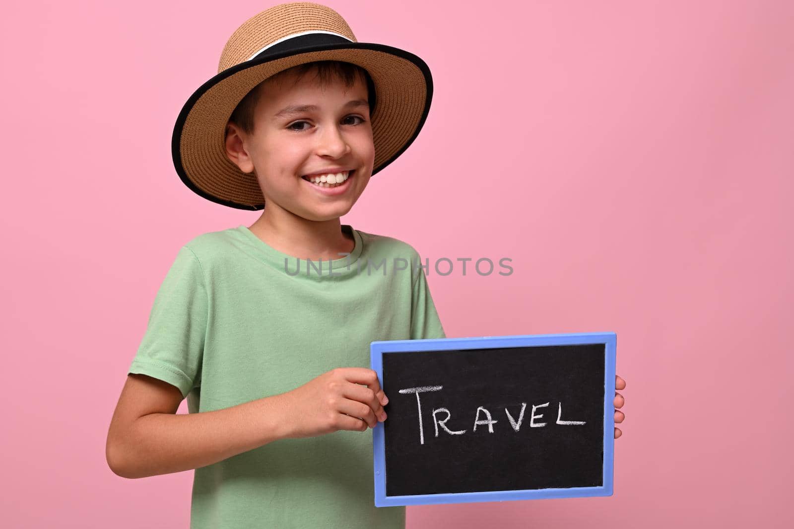 Adorable boy holding a board with travel lettering, smiling, looking at the camera. Pink background with copy space by artgf
