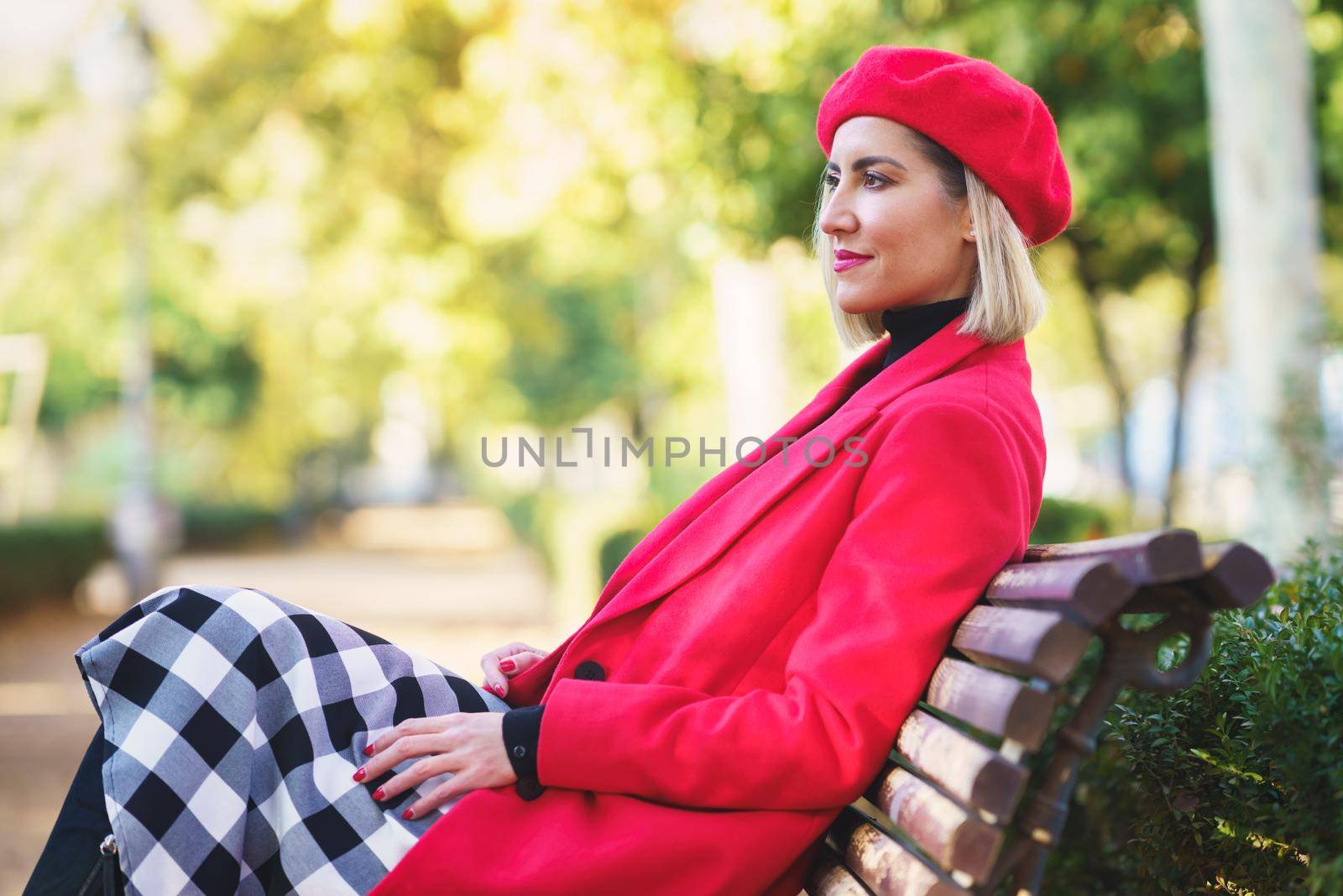 Middle-aged woman sitting on a bench in an urban park wearing red winter clothing. by javiindy