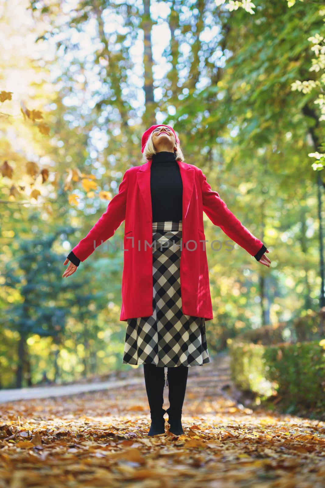 Happy woman opening her arms with her eyes closed in a park full of autumn leaves. Female wearing coat, skirt and beret outdoors.