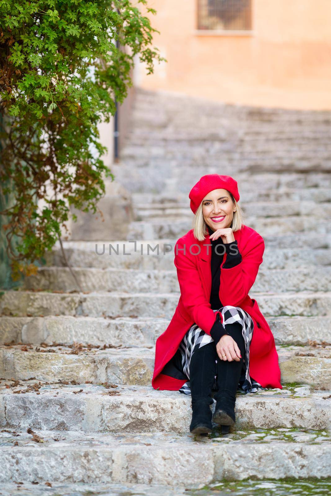 Woman wearing red winter clothes, sitting on steps in the Albaicin in Granada, Andalusia, Spain. Female wearing coat, skirt and beret outdoors.
