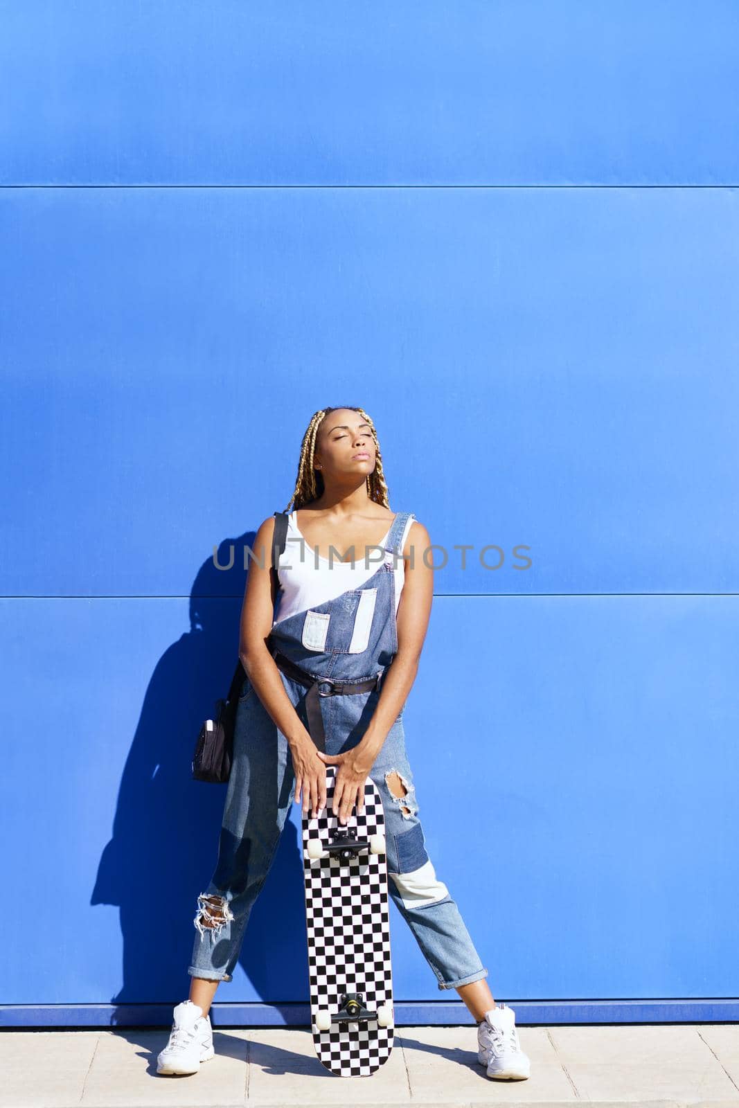 Black girl dressed casual, wtih a skateboard on blue wall background. by javiindy