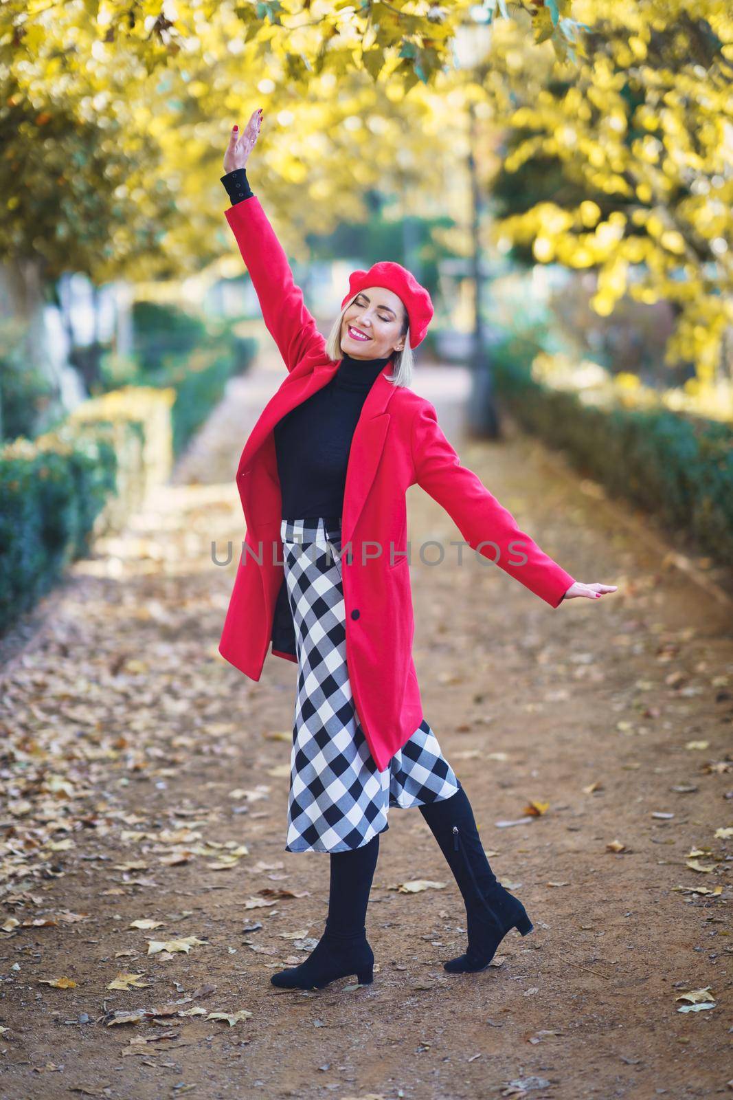 Happy woman wearing red winter clothes opening her arms with her eyes closed in a park full of autumn leaves. Female wearing coat, skirt and beret outdoors.