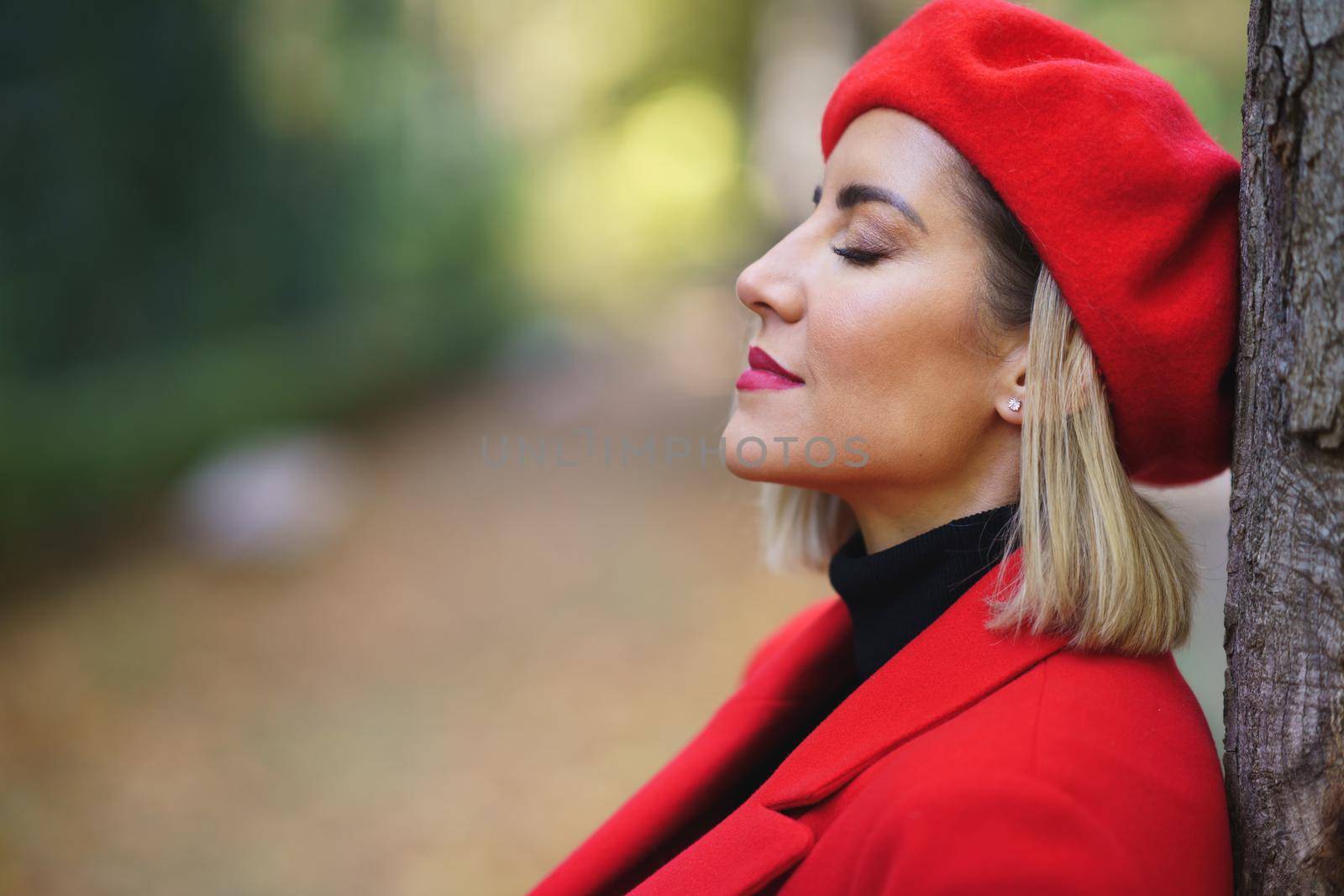 Close-up profile picture of a middle-aged woman leaning against a tree trunk with her eyes closed. Female in beret and red coat taking time to relax.