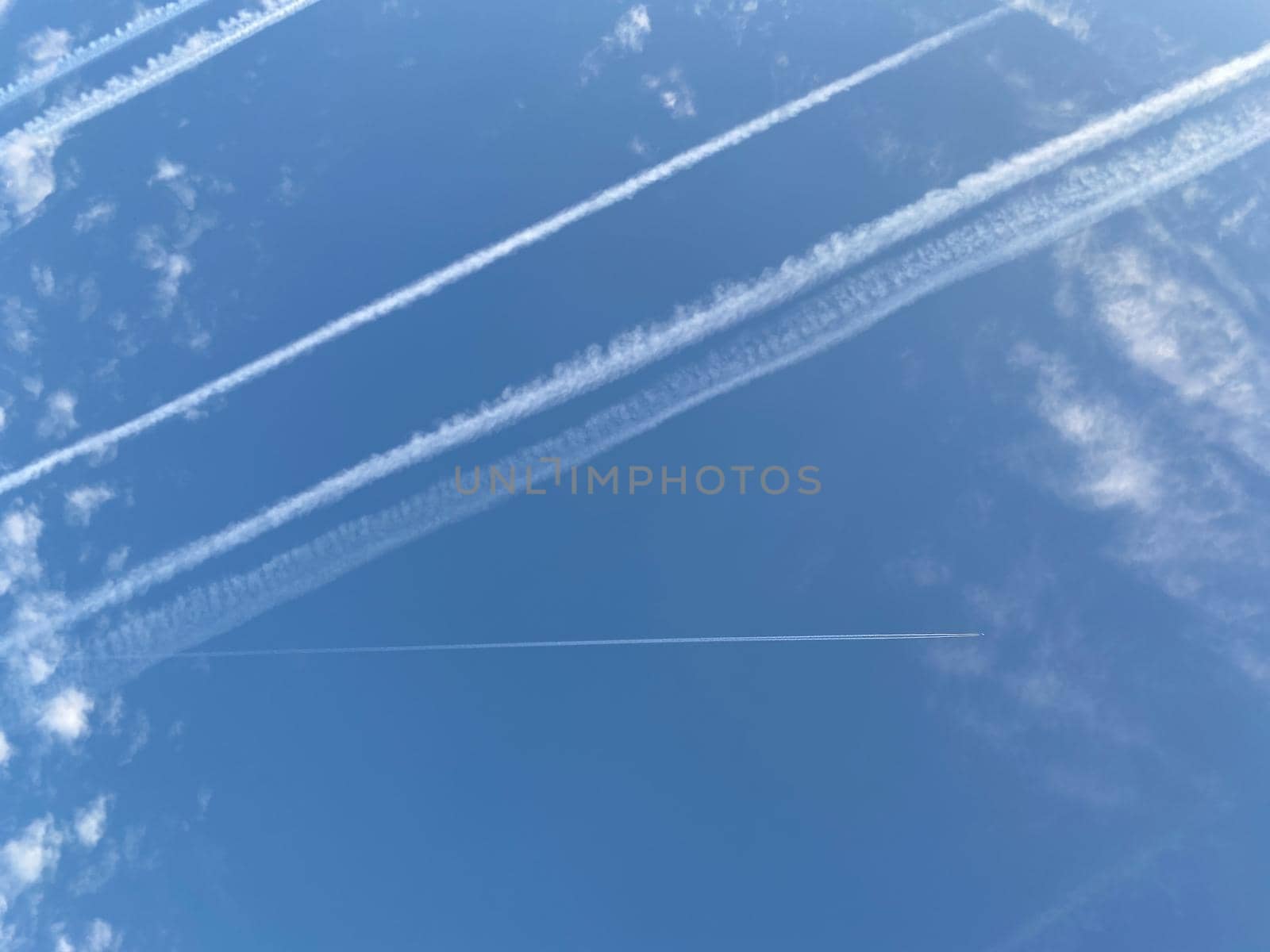 Airplane in the blue sky with clouds from below. High flying passenger plane with condensation trail. Jets flying overhead diagonally in sky with sunlight. Bottom view