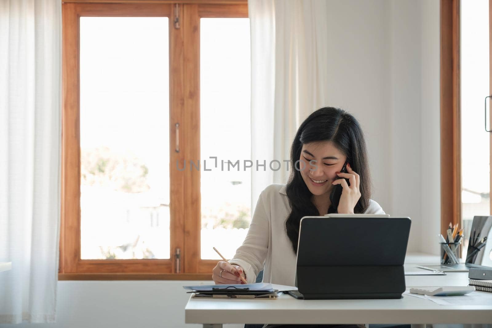 Happy business asian manageress working at her desk in the office taking a call on her mobile phone while writing notes on a notepad,