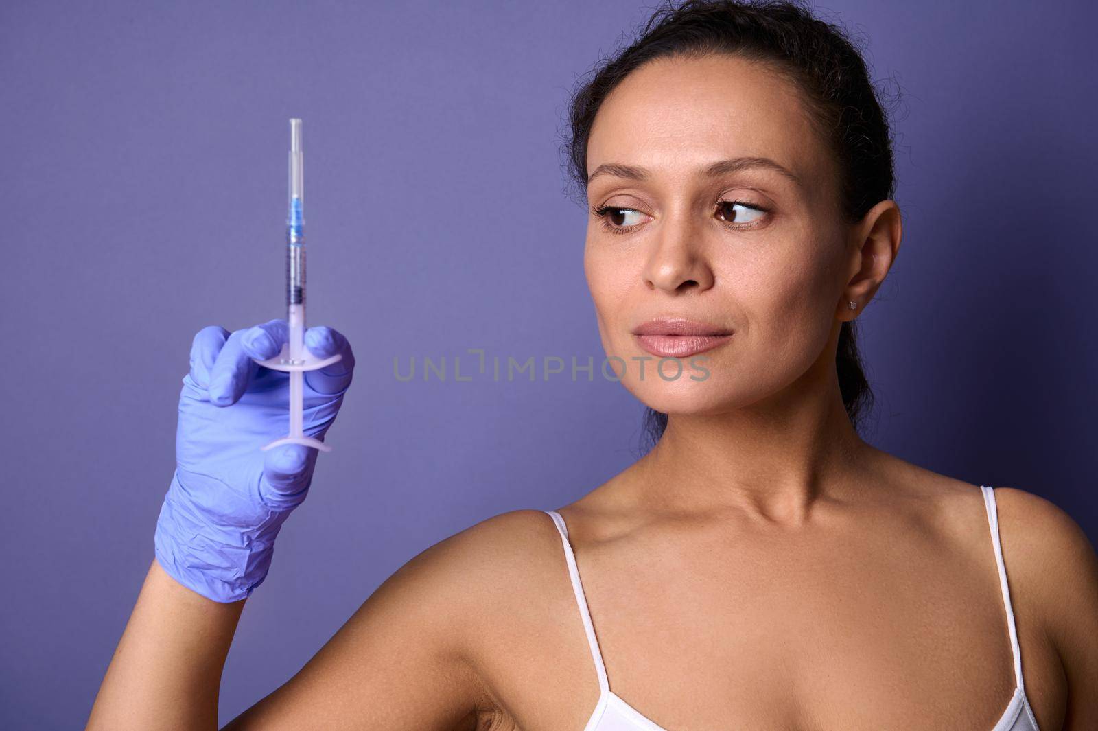 Natural beauty woman holding a syringe with rejuvenating injection in her hand, isolated on purple background with copy ad space. Cosmetology, aesthetic wellness spa concept. Healthcare and medicine
