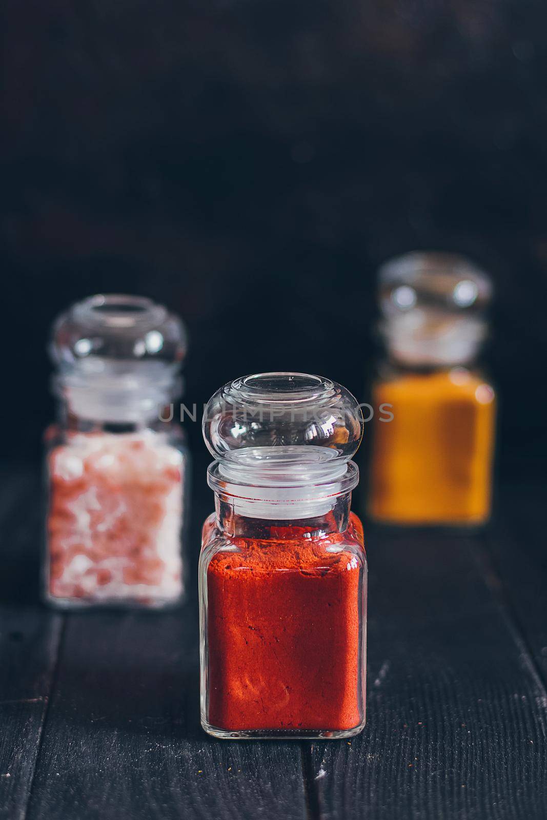 Bright aromatic spices in glass jars, assortments of spices: pink salt, paprika and turmeric on black stone background. by mmp1206