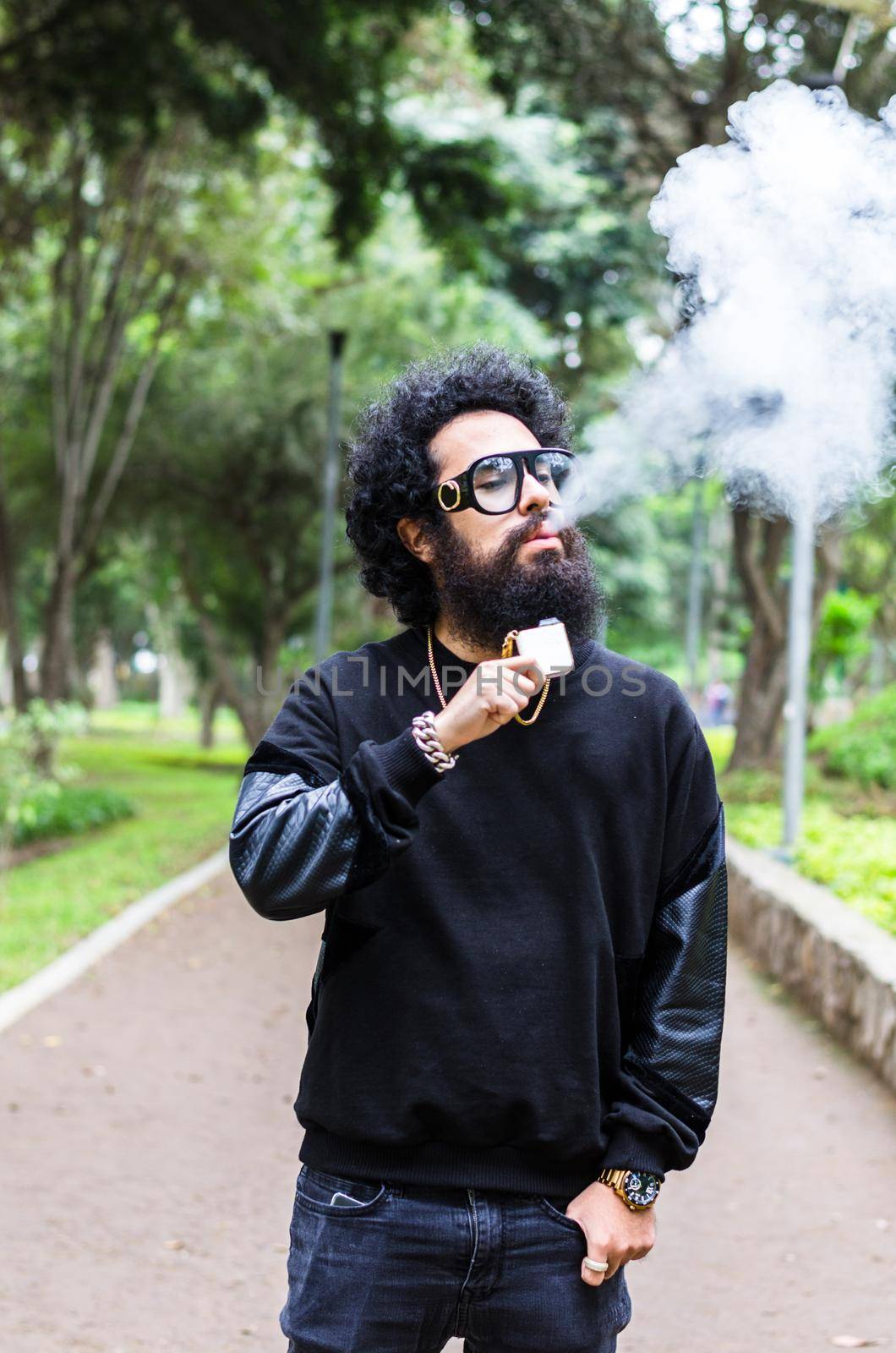 Vape. Young brutal man with large beard and fashionable haircut in sunglasses smoking an electronic cigarette in the city park. by Peruphotoart